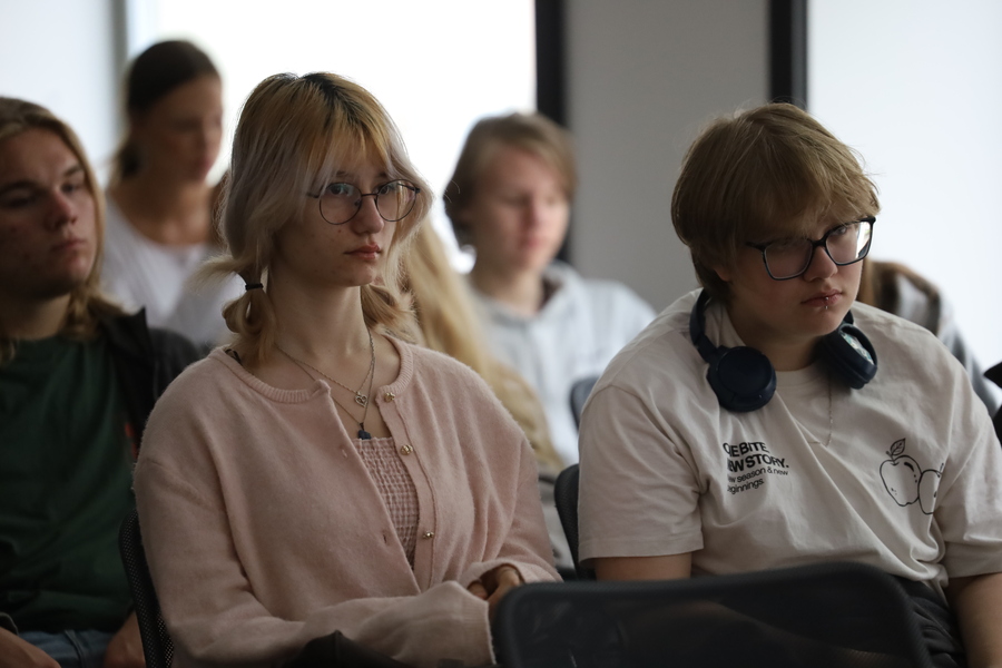 Grupa uczniów siedzących podczas zajęć edukacyjnych. Na pierwszym planie dwoje uczniów w okularach uważnie słucha prowadzącego; jedna z osób ma na szyi słuchawki.