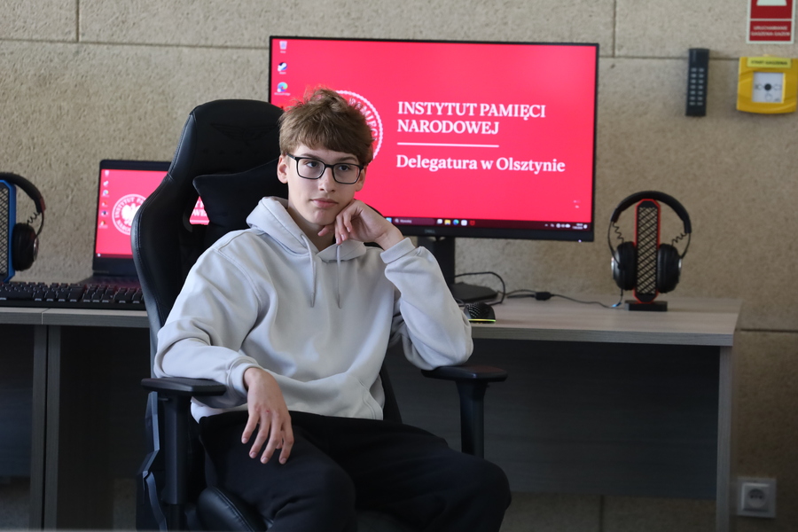 Uczeń w białej bluzie siedzi przy stanowisku komputerowym w Strefie Nowych Technologii. Za nim na monitorze widoczne jest logo Instytutu Pamięci Narodowej – Delegatury w Olsztynie.
