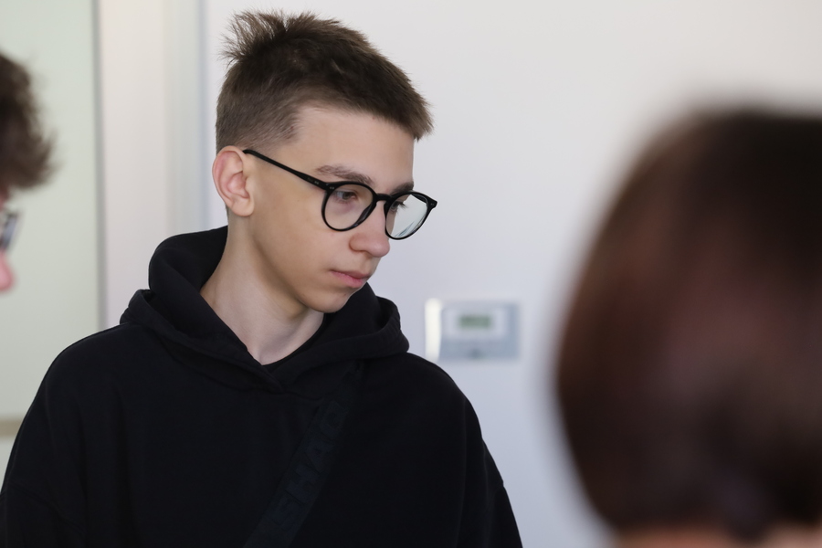 Uczeń w czarnej bluzie i okularach podczas zajęć edukacyjnych. Stoi wśród pozostałych uczestników i uważnie słucha osoby znajdującej się poza kadrem.