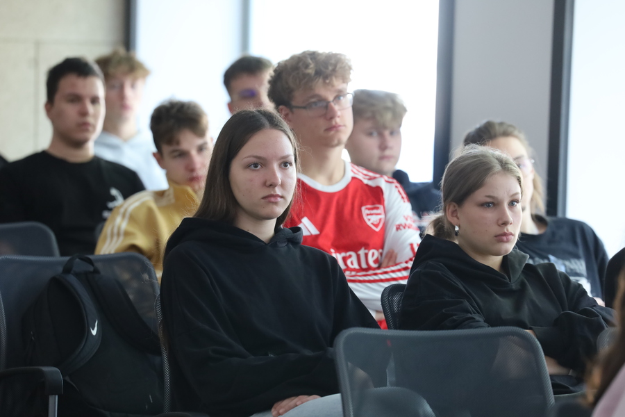Grupa uczniów siedzących w sali podczas zajęć edukacyjnych. Młodzież uważnie słucha prowadzącego znajdującego się poza kadrem.