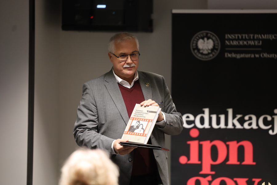 Dr Waldemar Brenda prezentuje uczestnikom książkę „Filmowcy w rzeczywistości politycznej Polski Ludowej”. Za nim znajduje się baner IPN.