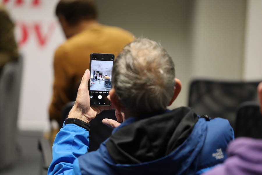 Uczestnik spotkania w niebieskiej kurtce fotografuje telefonem komórkowym wydarzenie odbywające się w Delegaturze IPN w Olsztynie.