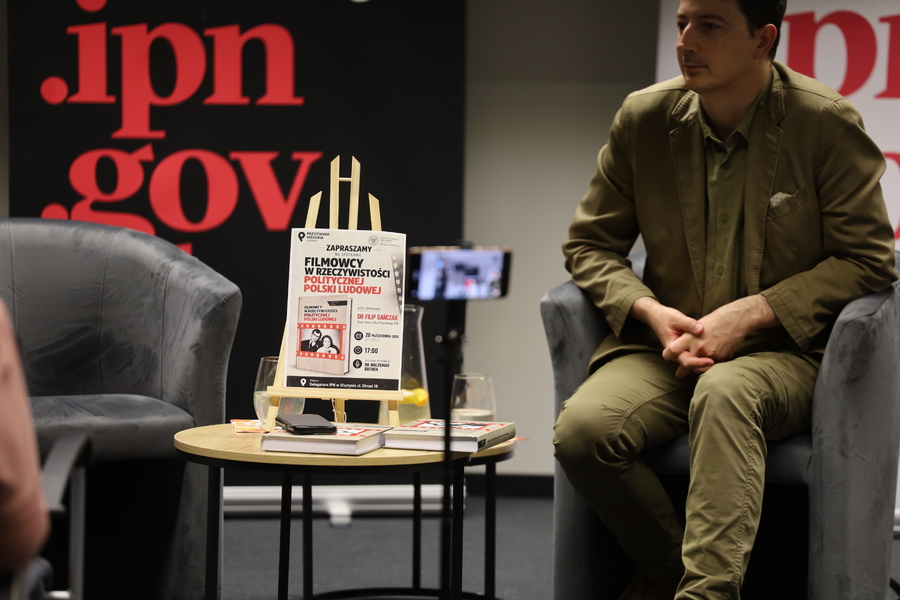 Dr Filip Gańczak siedzi w fotelu obok stolika, na którym znajdują się książki i plakat zapowiadający spotkanie poświęcone filmowcom w PRL.