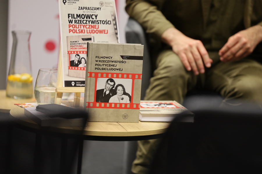 Na stoliku stoi książka „Filmowcy w rzeczywistości politycznej Polski Ludowej”. Za nią widoczny plakat wydarzenia, a w tle siedzący dr Filip Gańczak.