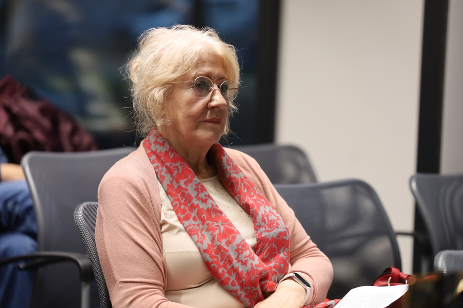 Uczestniczka spotkania w okularach, różowym swetrze i czerwono-szarym szaliku siedzi na krześle, trzymając kartki.