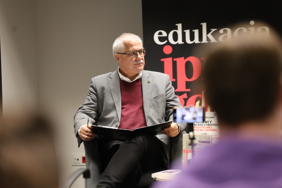 Dr Waldemar Brenda siedzi w fotelu z otwartą teczką na kolanach, prowadząc spotkanie z cyklu „Przystanek Historia”.